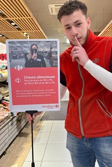 Un employé de Carrefour met un doigt devant sa bouche à côté d'un panneau annonçant le dispositif de l'heure silencieuse