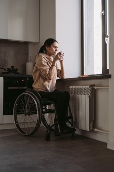 Une femme en chaise roulante boit un thé dans sa cuisine en regardant par la fenêtre