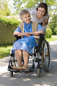 Une aide à domicile accompagne une personne âgée souriante avec son fauteuil roulant